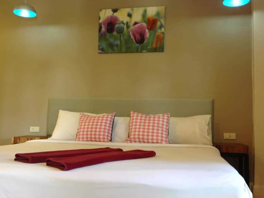 Un lit bien fait, avec du linge de lit blanc et des coussins à carreaux rouges, une serviette rouge posée sur le lit et un tableau de fleurs accroché au mur.