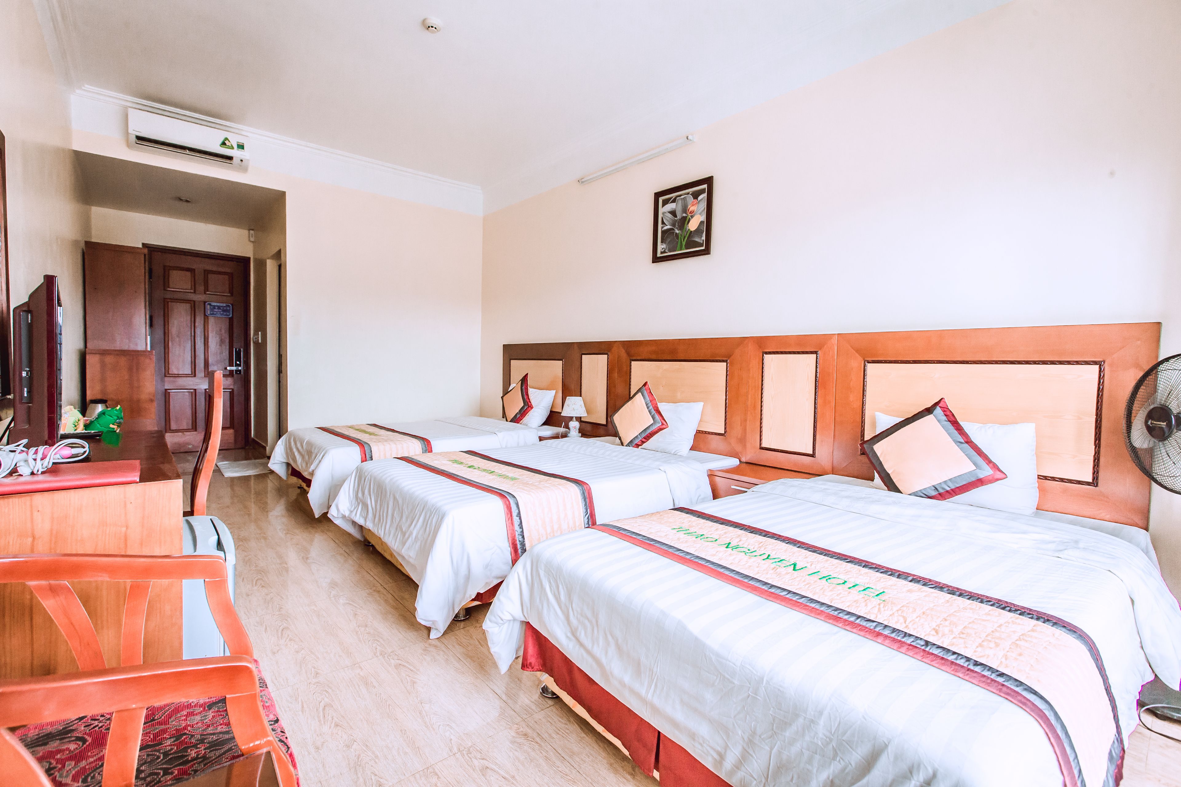 A hotel room with three beds, a desk with a chair, and a fan.