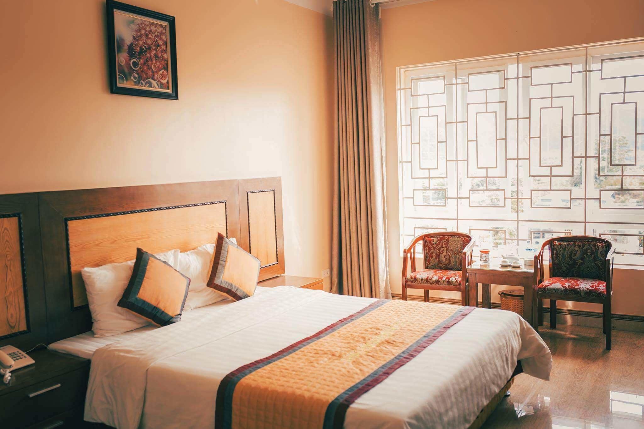 A hotel room with a large bed, a wooden headboard, two chairs, a table, and a window with a geometric pattern.