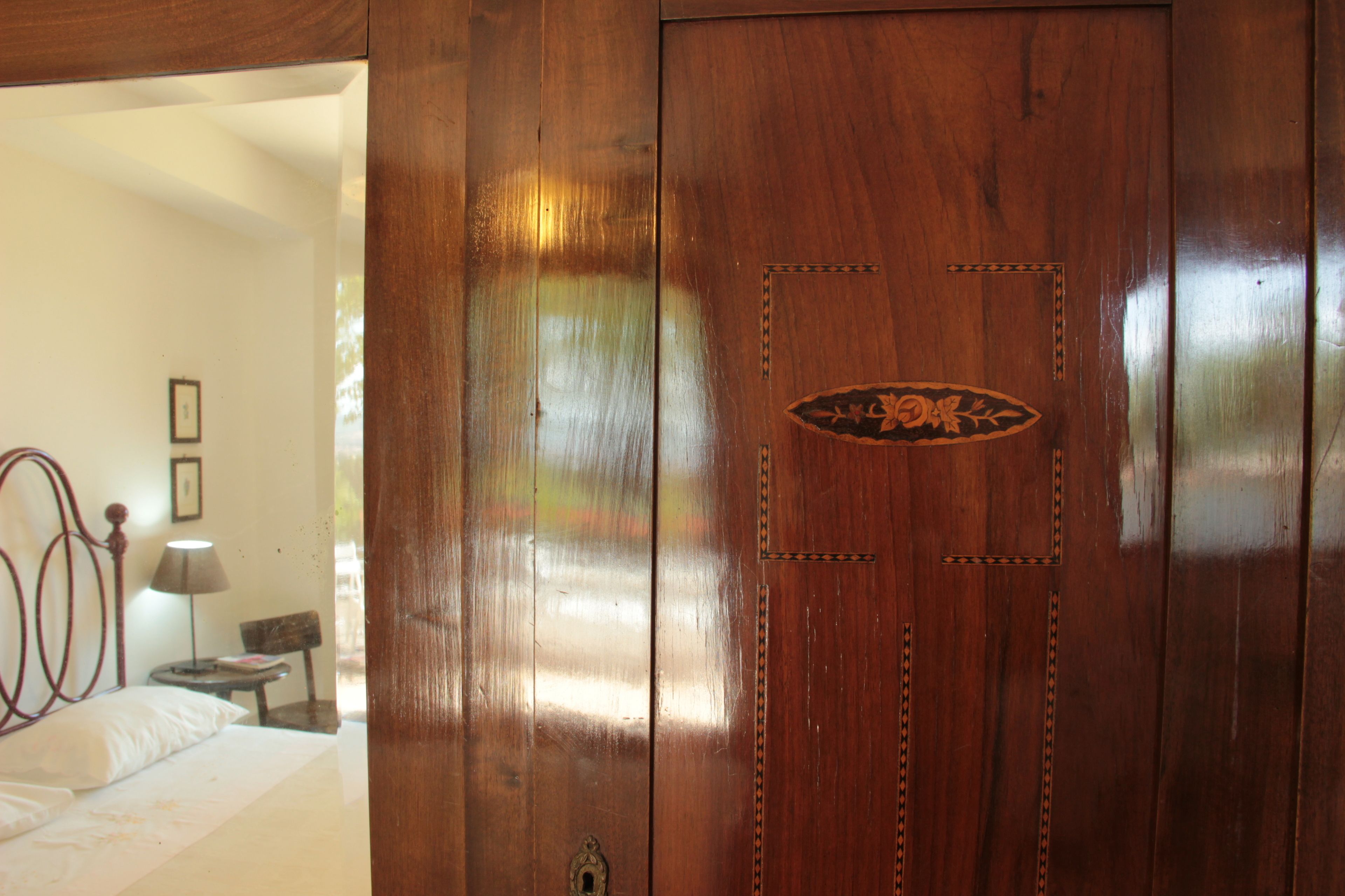 A wooden door with a decorative emblem, a bed with a white bedspread, a nightstand with a lamp, and a chair in a room with white walls.