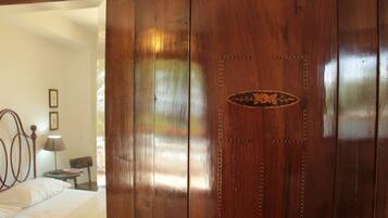 A wooden door with a decorative emblem, a bed with a white bedspread, a nightstand with a lamp, and a chair in a room with white walls.