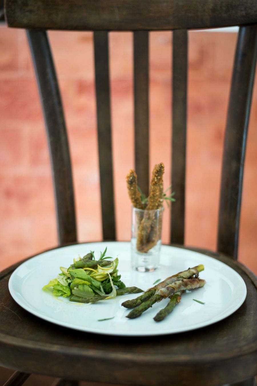 A white plate with asparagus and a small glass containing a plant on a wooden chair.