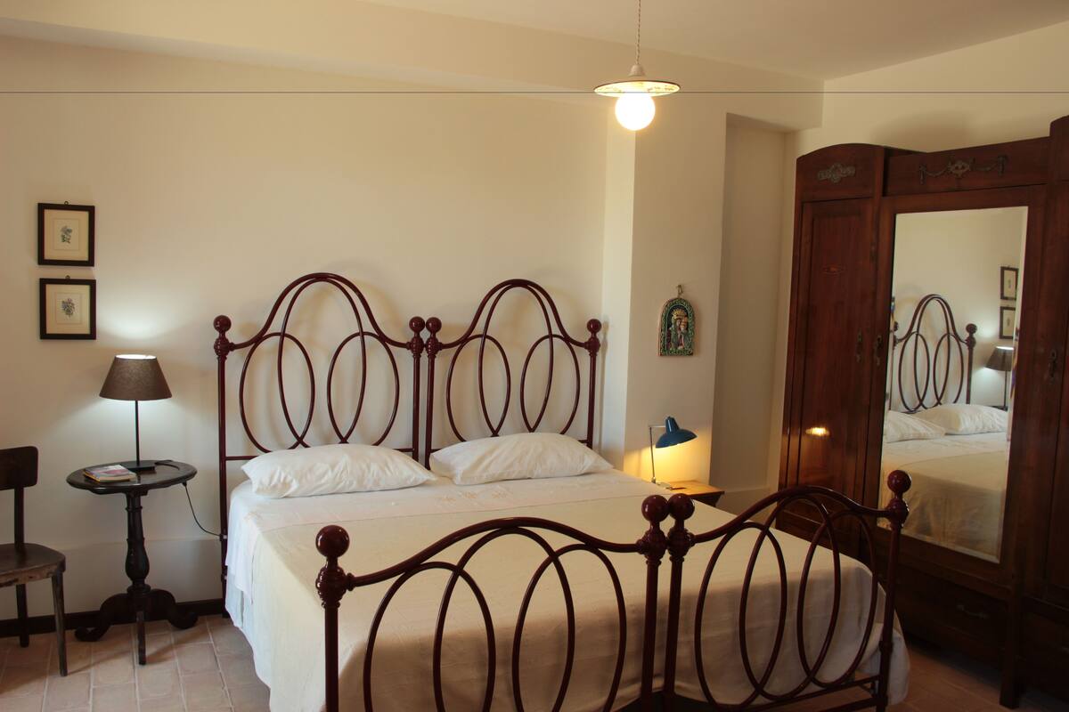 A bedroom with a wooden four-poster bed, a bedside table with a lamp, a chair, and a wardrobe.