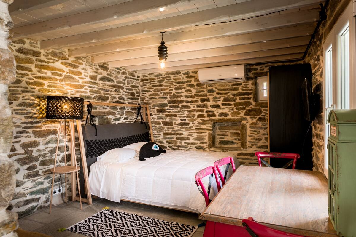A bedroom with a stone wall, a bed with a white bedspread, a wooden ladder, a red chair, and a patterned rug.