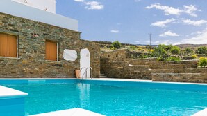 A swimming pool with stone walls, a white building with wooden doors, and a clear blue sky. - imarkellis boutique villas (Tinos)