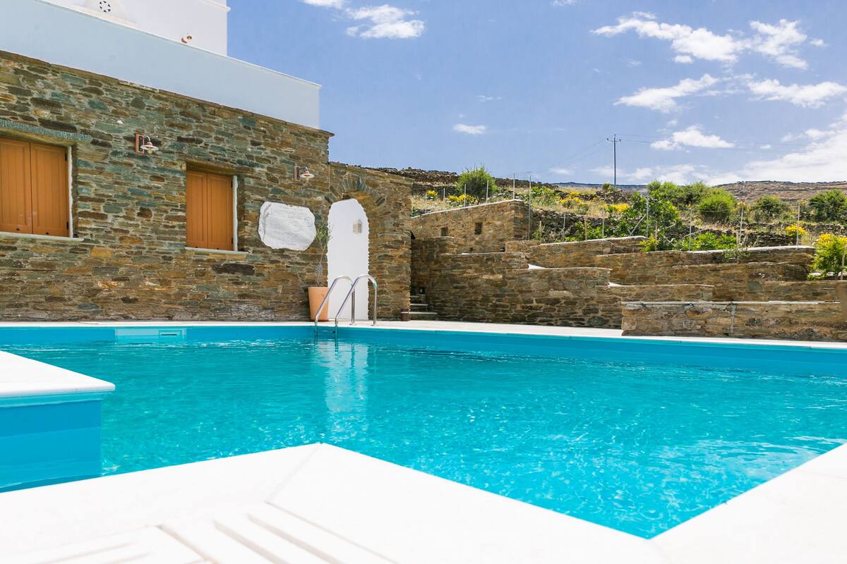 A swimming pool with stone walls, a white building with wooden doors, and a clear blue sky.