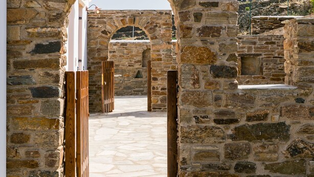 An arched stone doorway leading to a courtyard with stone flooring and a white building in the background. - imarkellis boutique villas (Tinos)