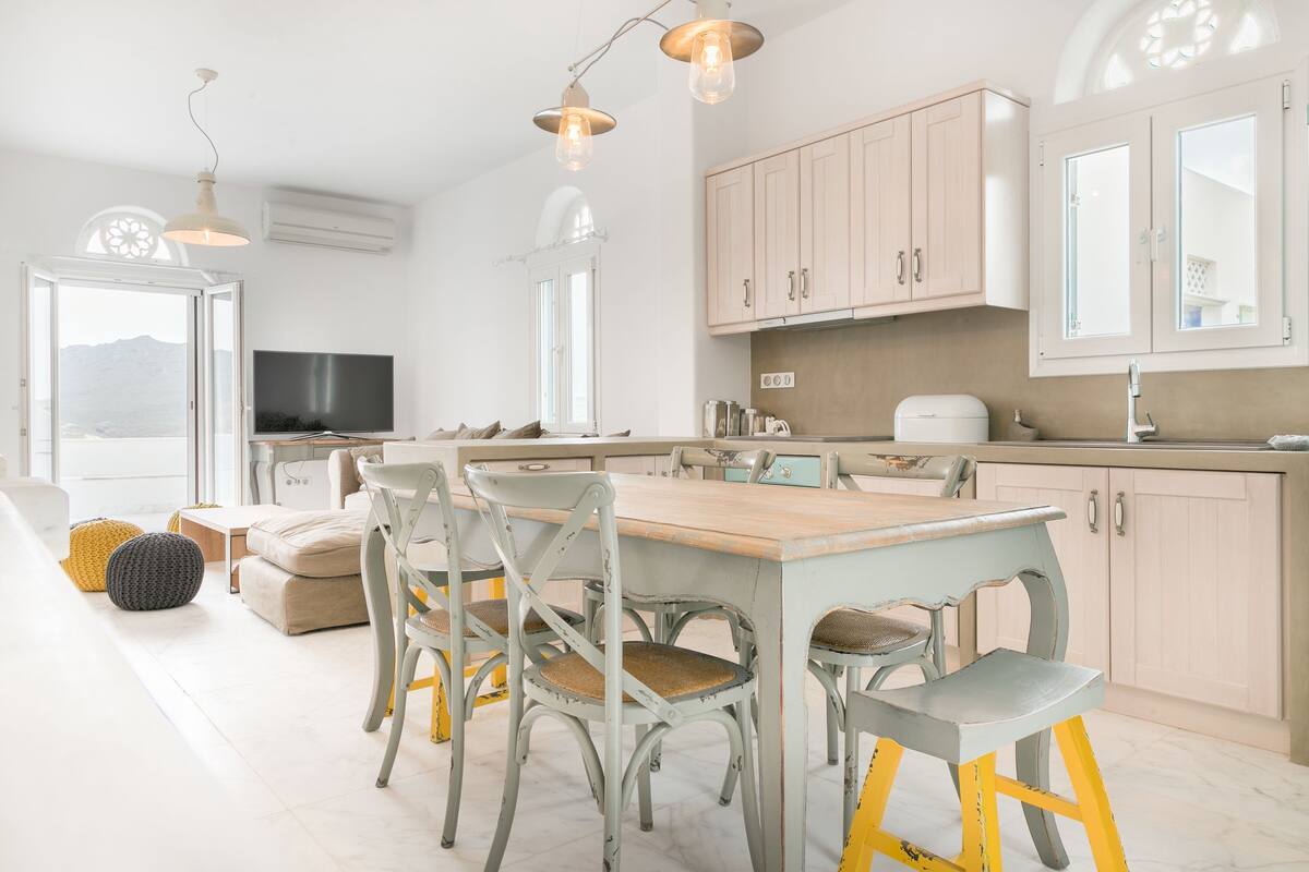 A modern kitchen with a dining area, featuring a wooden table, grey chairs, and a large window.