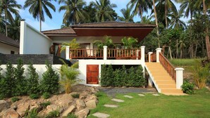 A two-story house with a wooden balcony, a stone pathway leading to the entrance, and a lush garden with palm trees. - Tropical Season Villa Resort (Koh Samui)