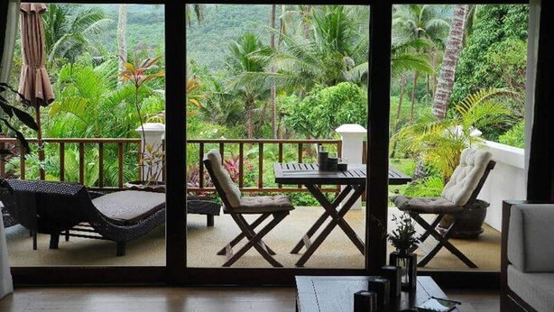 A balcony with wicker furniture, a table, and chairs, overlooking a lush garden with palm trees. - Tropical Season Villa Resort (Koh Samui)