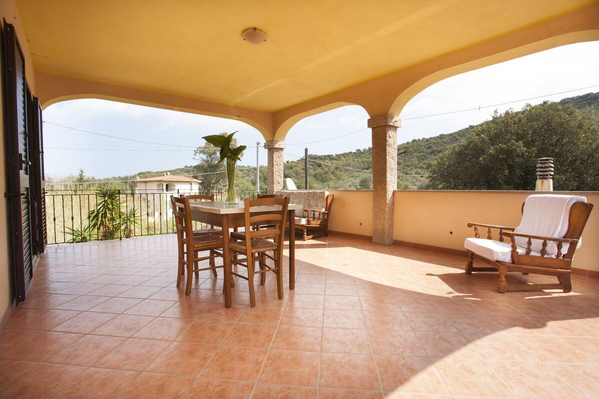 Une terrasse extérieure couverte, avec une table et des chaises, un banc et une vue sur un paysage vallonné.