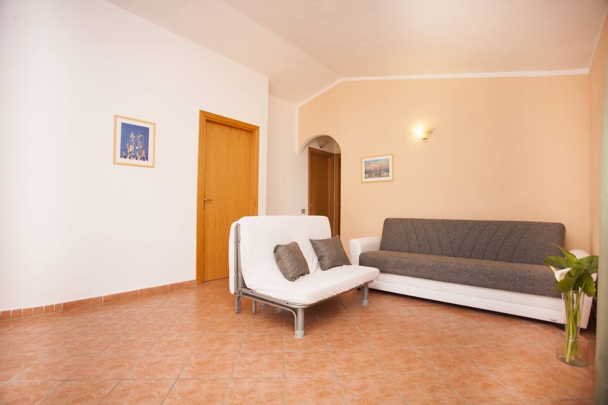 Une pièce avec un canapé blanc, un petit fauteuil blanc, une porte en bois et un luminaire fixé au mur.