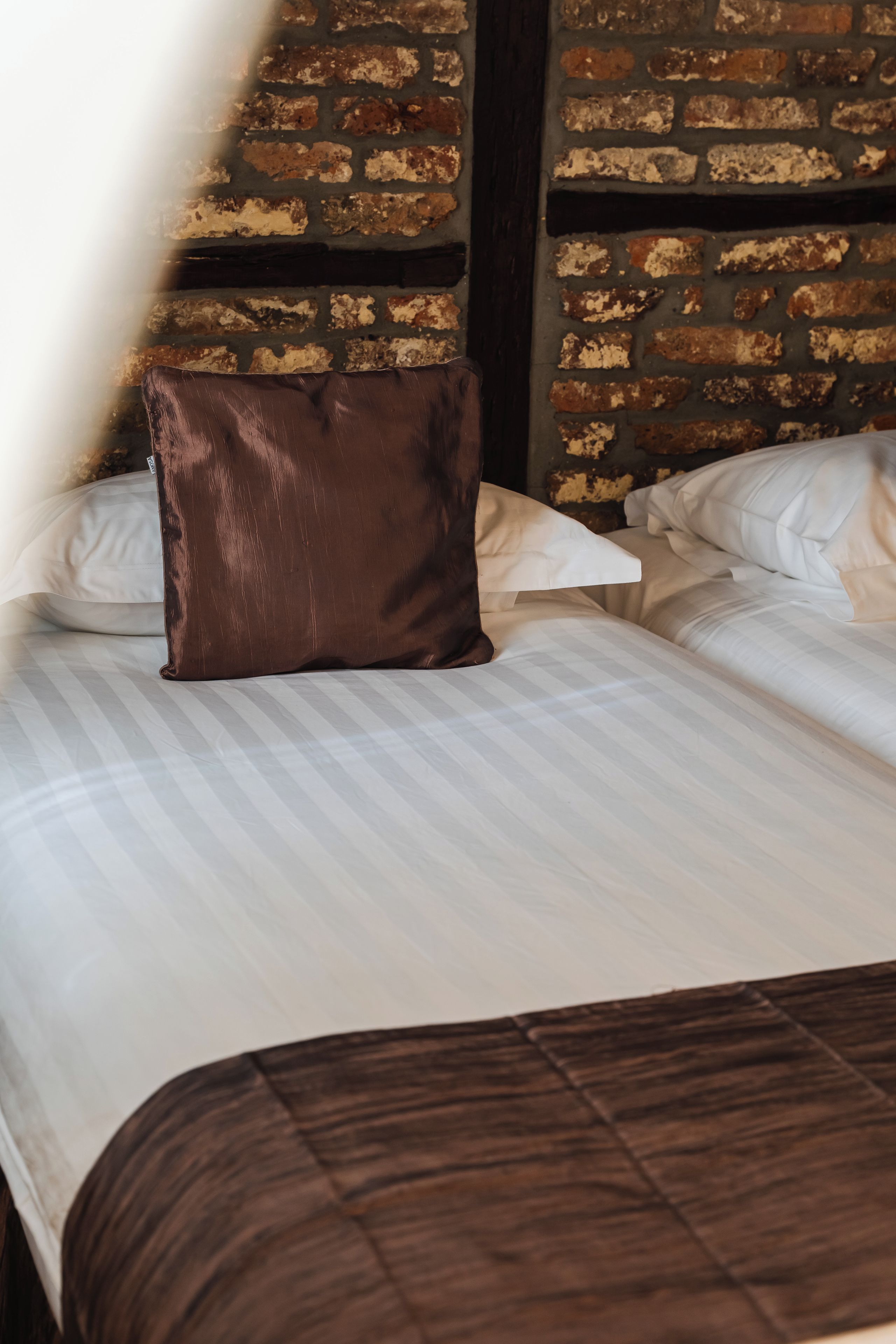 Una cama con ropa blanca y un almohadón marrón, frente a una pared de ladrillo.