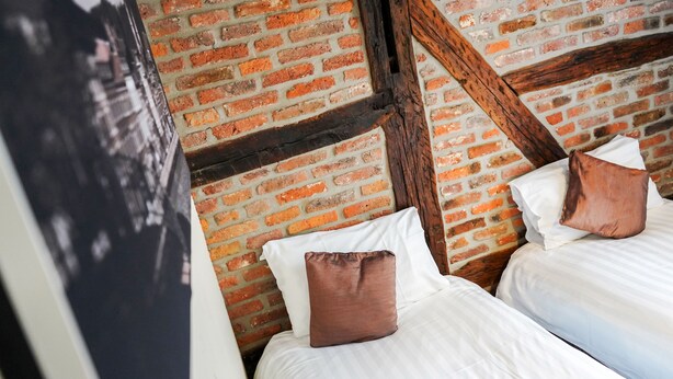 Una habitación con pared de ladrillo y vigas de madera a la vista. Hay dos camas con ropa blanca y almohadas marrones.
