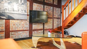 Una sala de estar moderna con una mesa de centro de vidrio, una pared de ladrillo, una escalera de madera y un televisor de pantalla plana montado en la pared.