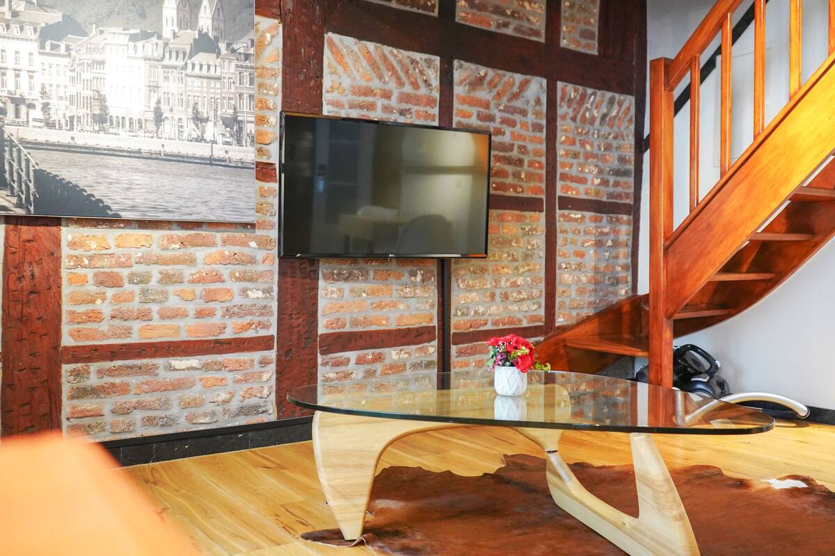 Una sala de estar moderna con una mesa de centro de vidrio, una pared de ladrillo, una escalera de madera y un televisor de pantalla plana montado en la pared.