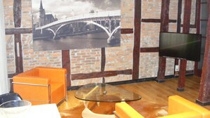 Una habitación con una televisión grande montada en una pared de ladrillo, sillas naranjas, una mesa de cristal y un cuadro con una imagen de un puente.