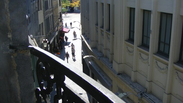 Une vue depuis un balcon donnant sur une rue pavée, avec des piétons et des bâtiments.