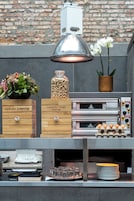 Una zona de cocina con fondo de pared de ladrillo, una lámpara colgante, una caja de madera con la etiqueta 'FORREST FRUIT SMOOTHIES', un tarro de cacahuates, una orquídea en maceta y una pila de platos.