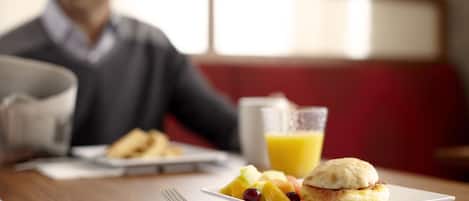 A breakfast table with a plate of fruit, a sandwich, a glass of orange juice, and a cup.