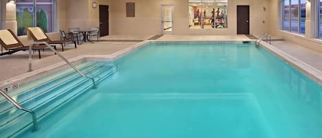 A large indoor swimming pool with lounge chairs and a table, surrounded by walls with windows and a door.