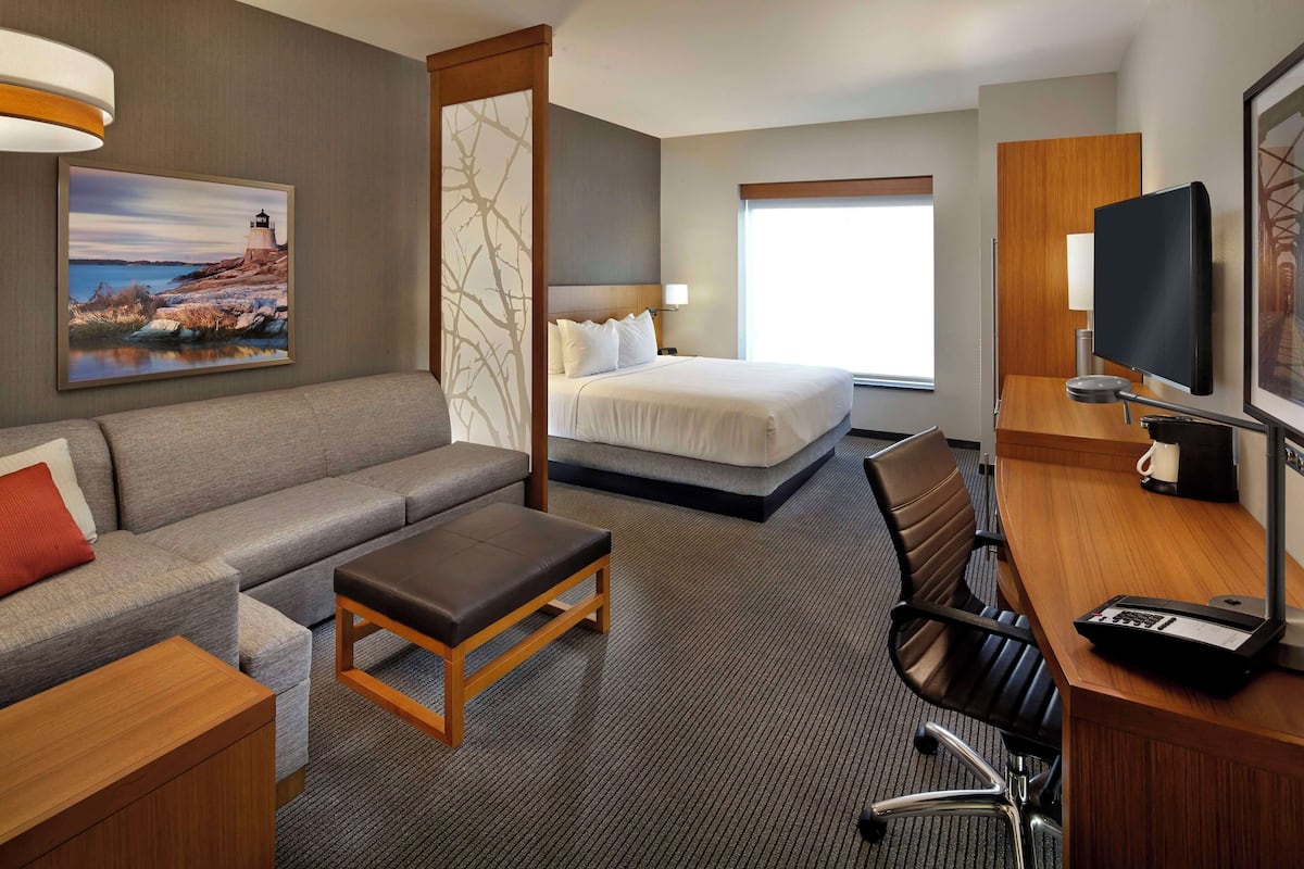Ein Hotelzimmer mit einem Bett, einem Sofa, einem Schreibtisch, einem Sessel, einem Fernseher und einem Gemälde eines Leuchtturms.