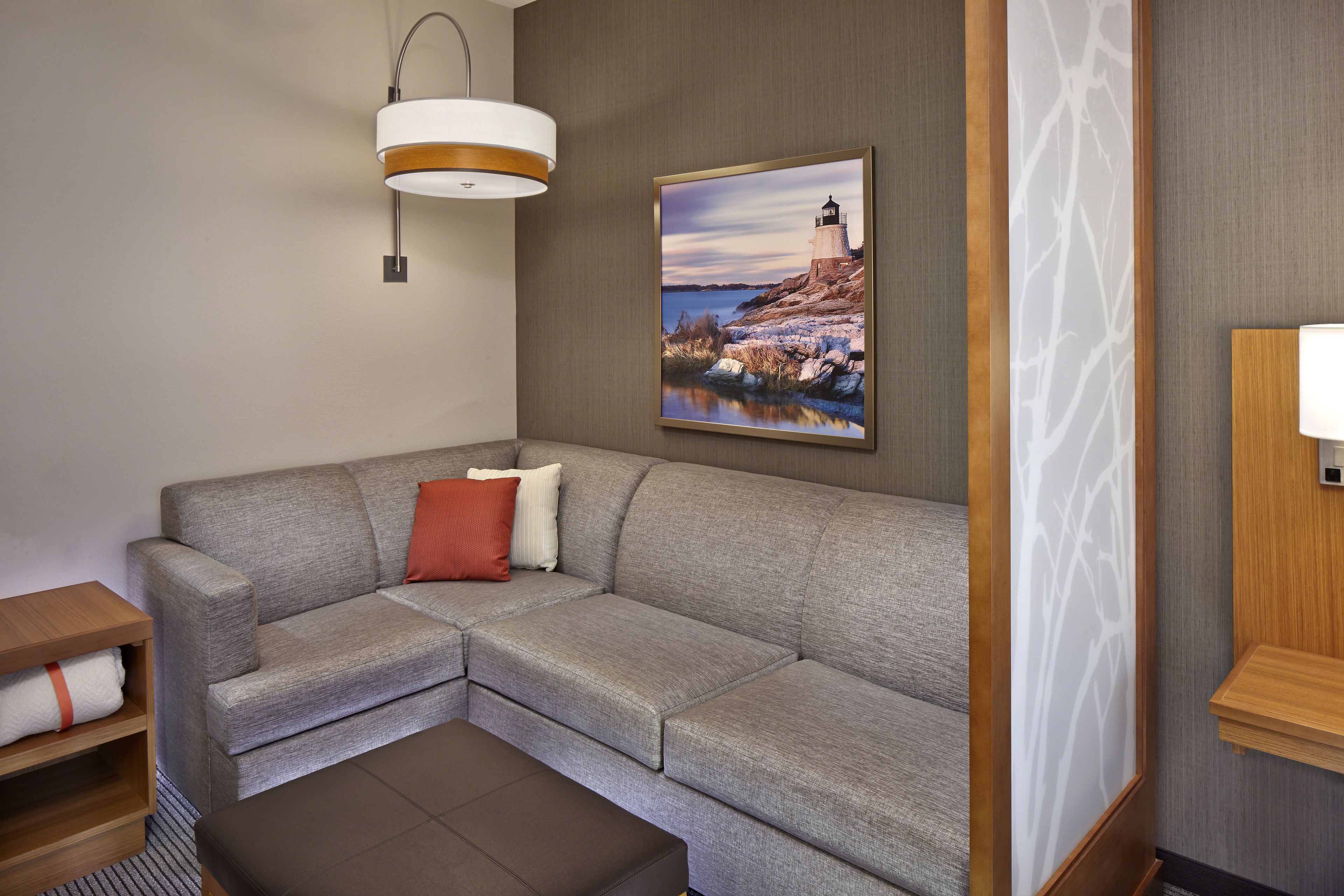 Un salon moderne avec un canapé d’angle gris, une table d’appoint en bois, un pouf marron, une lampe fixée au mur et un tableau encadré représentant un phare.