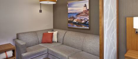 A modern living room with a grey sectional sofa, a wooden side table, a brown ottoman, a wall-mounted lamp, and a framed picture of a lighthouse.