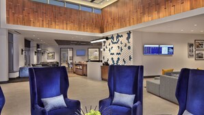 Un hall d’entrée moderne avec des fauteuils bleus, une table ronde en bois sur laquelle sont disposées des pommes, et un comptoir d’accueil équipé d’écrans.