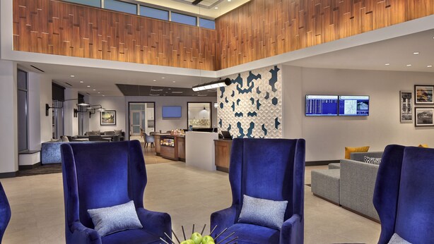 Eine moderne Lobby mit blauen Sesseln, einem runden Holztisch mit Äpfeln und einem Empfangsbereich mit Monitoren.