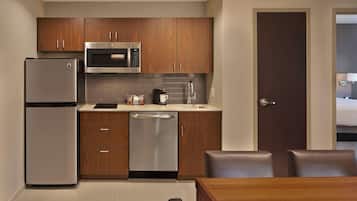 A compact kitchen with wooden cabinets, a stainless steel refrigerator, a microwave, and a dishwasher. There is a dining table with chairs and a door leading to another room.
