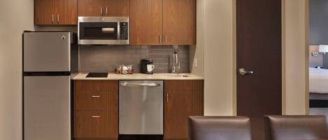 A compact kitchen with wooden cabinets, a stainless steel refrigerator, a microwave, and a dishwasher. There is a dining table with chairs and a door leading to another room.
