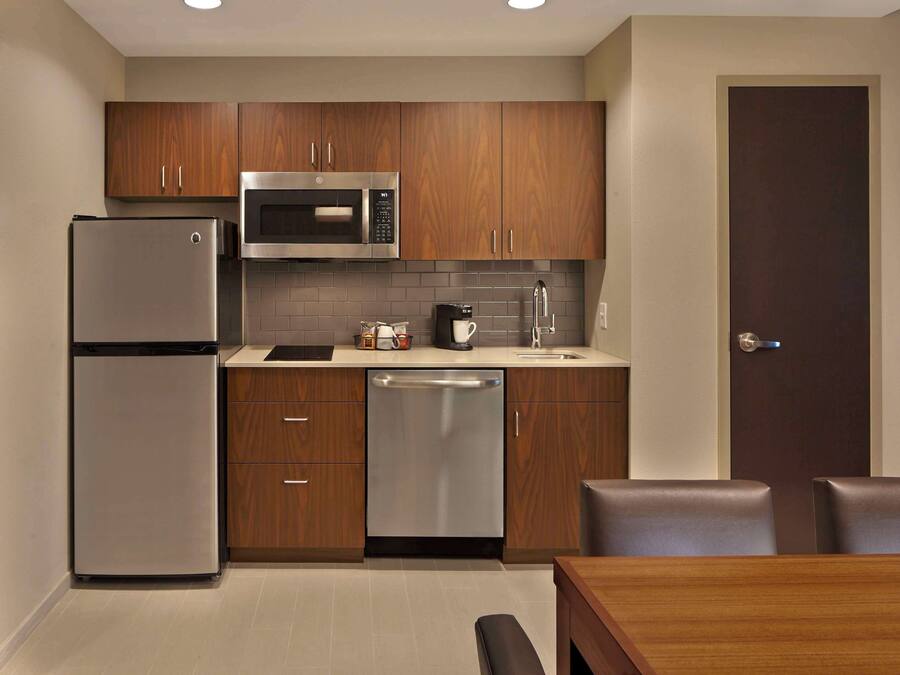 A compact kitchen with wooden cabinets, a stainless steel refrigerator, a microwave, and a dishwasher. There is a dining table with chairs and a door leading to another room.
