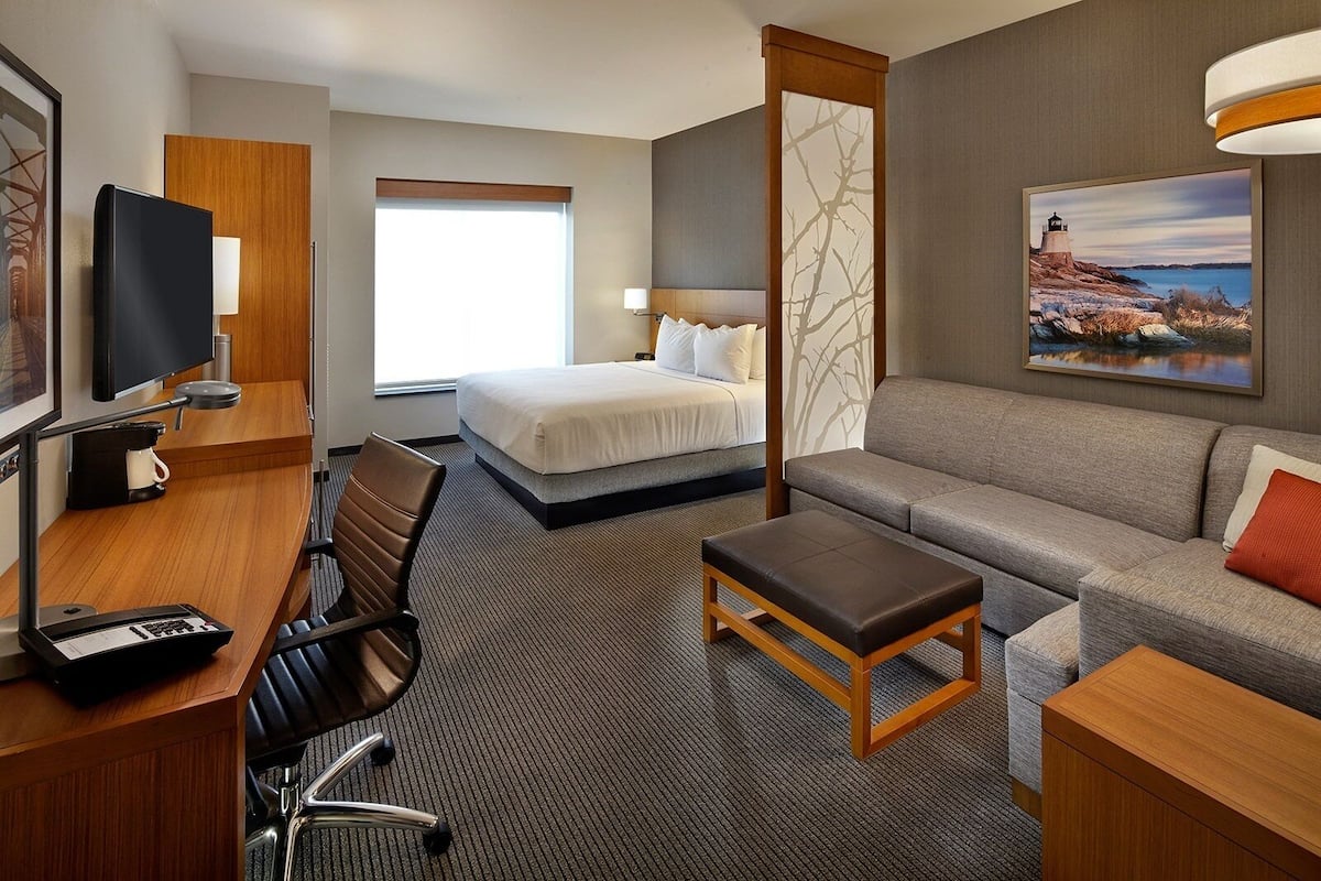 Ein Hotelzimmer mit einem Bett, einem Schlafsofa, einem Schreibtisch mit Stuhl, einem Fernseher und einem Gemälde eines Leuchtturms.