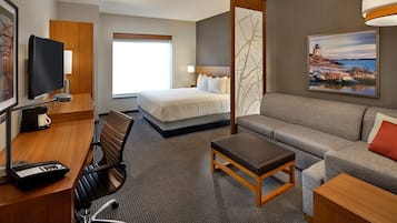 A hotel room with a bed, a sofa, a desk with a chair, a TV, and a painting of a lighthouse.