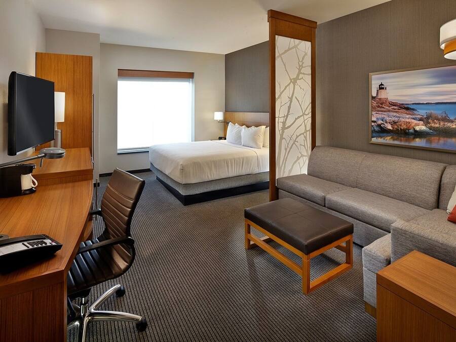 A hotel room with a bed, a sofa, a desk with a chair, a TV, and a painting of a lighthouse.