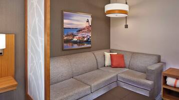 A modern living room with a grey sectional sofa, a small wooden side table, a wall-mounted lamp, and a framed picture of a lighthouse.