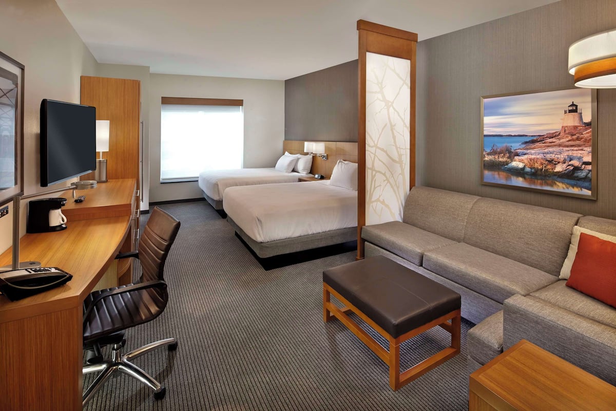 Ein Hotelzimmer mit zwei Betten, einem Schlafsofa, einem Schreibtisch mit Fernseher und einem Bild an der Wand.
