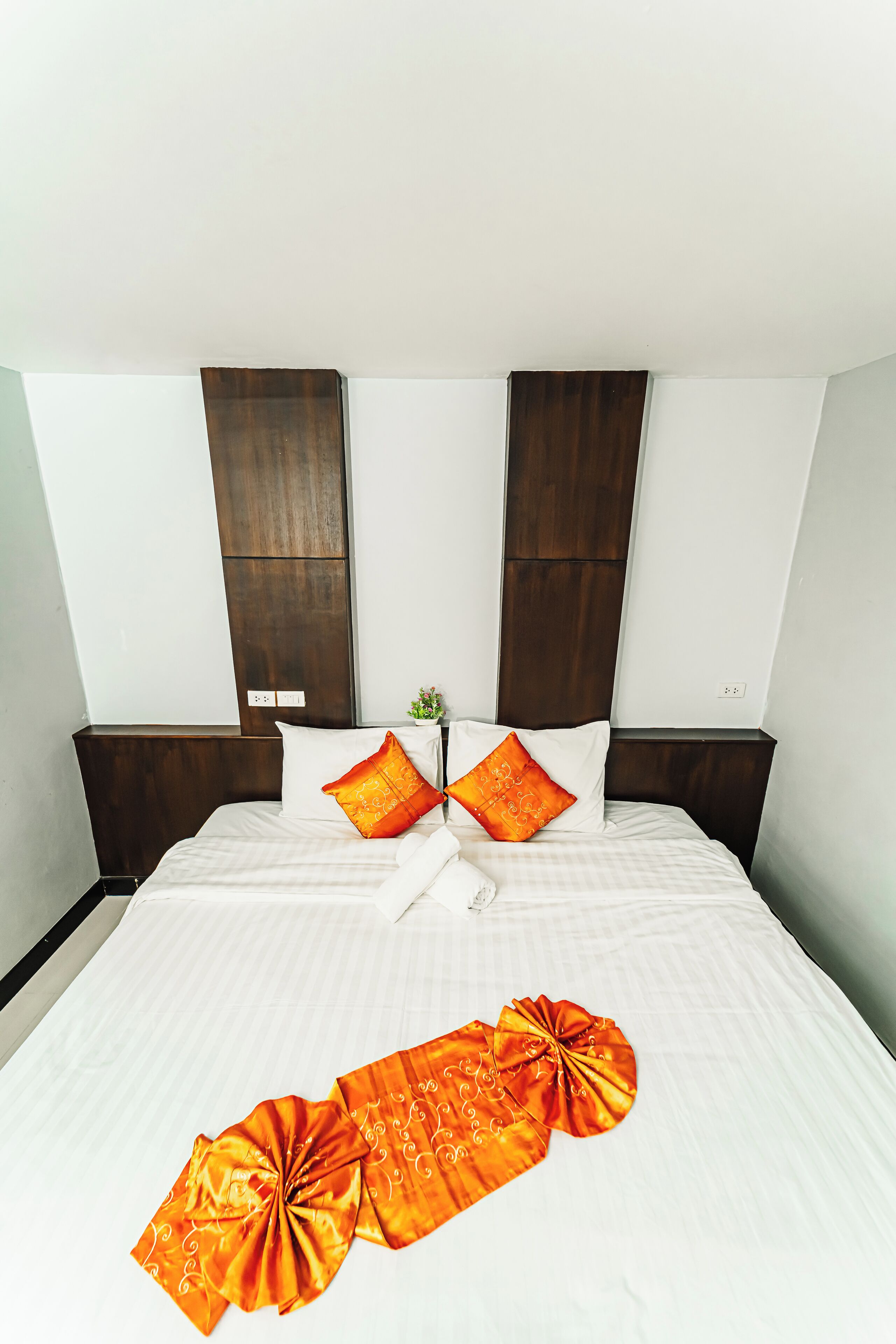 A neatly made bed with white linens and two orange pillows, a wooden headboard, and a small plant on the nightstand.