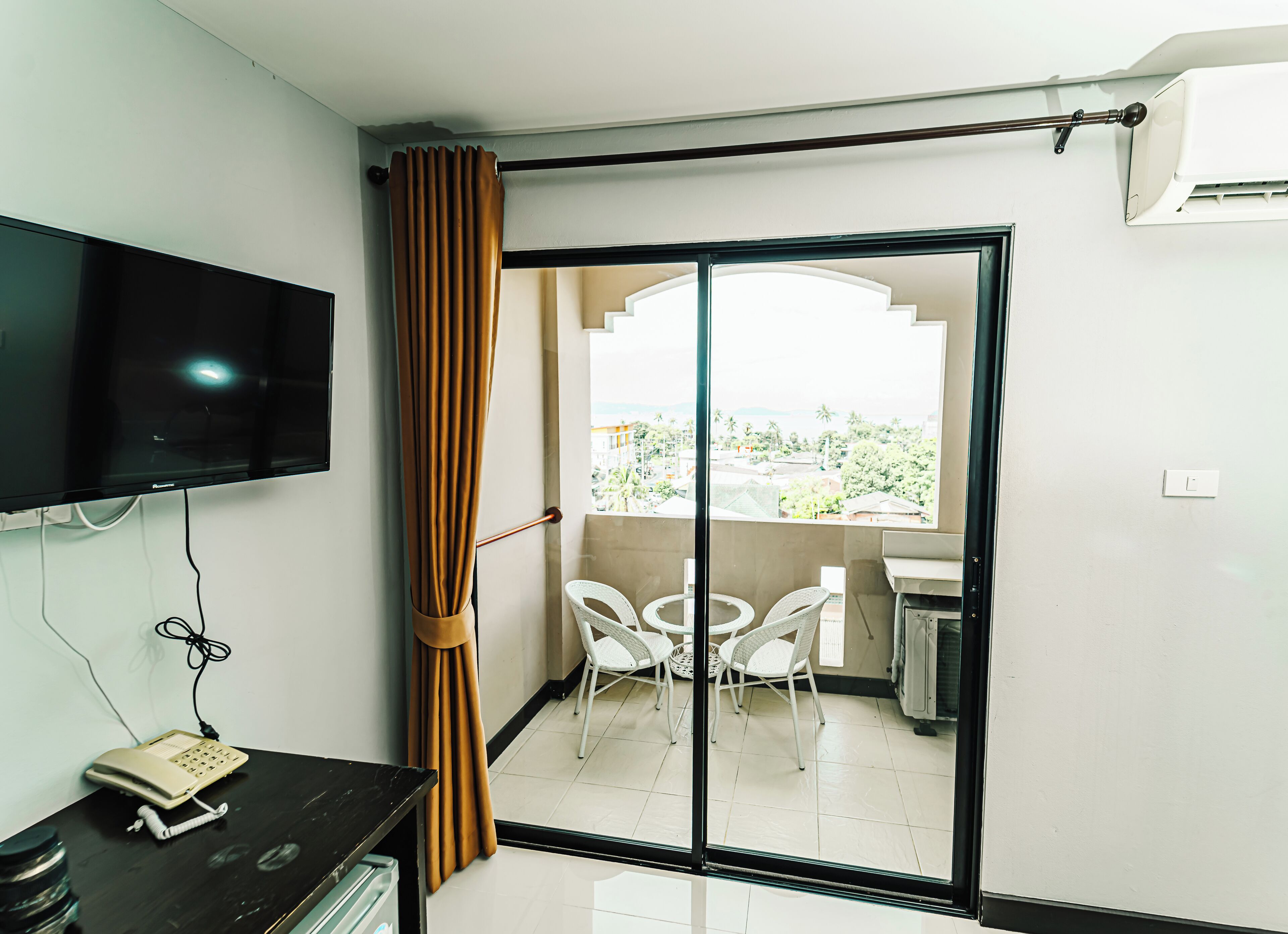 A room with a sliding glass door leading to a balcony with white chairs and a table. A television is mounted on the wall, and a telephone is on a desk.