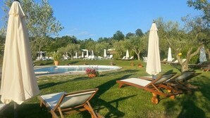Zona junto a la piscina con sillas de descanso y sombrillas, rodeada de follaje y árboles.