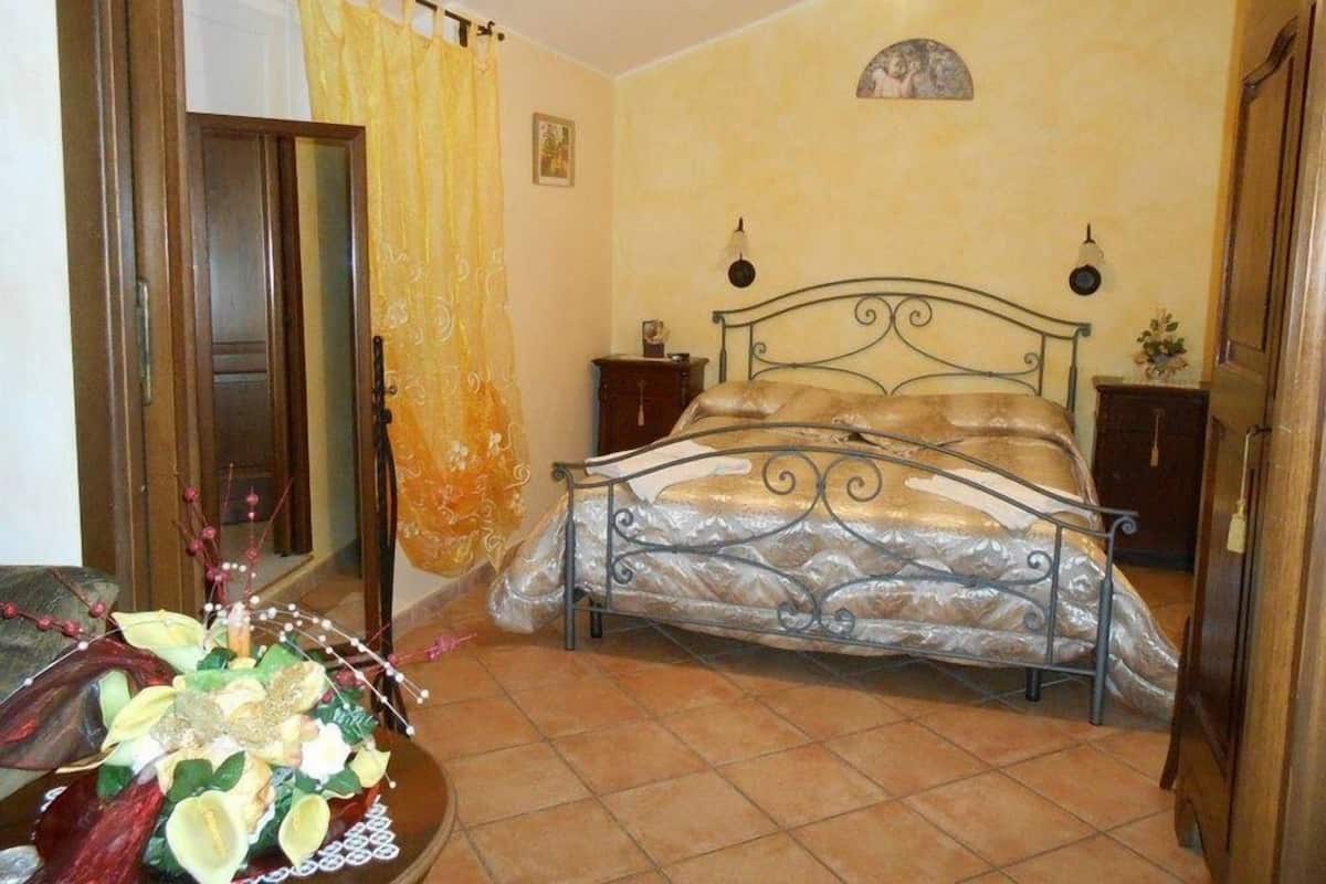 Un dormitori amb un llit, una porta de fusta, una cortina, una làmpada fixa a la paret i una petita taula de nit.