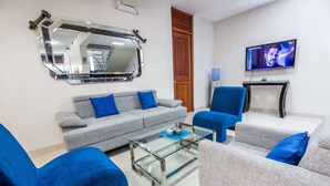 A modern living room with a grey sofa, blue armchairs, and a glass coffee table. A large mirror and a flat-screen TV are mounted on the wall. - Hotel El Sol (Piura)