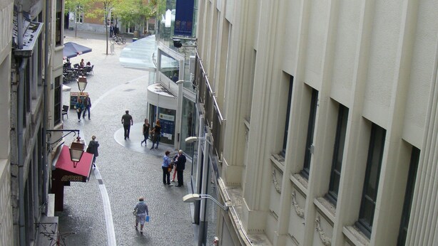 Una calle adoquinada flanqueada por edificios, un cartel que indica Galerie St. Lambert, y gente caminando.