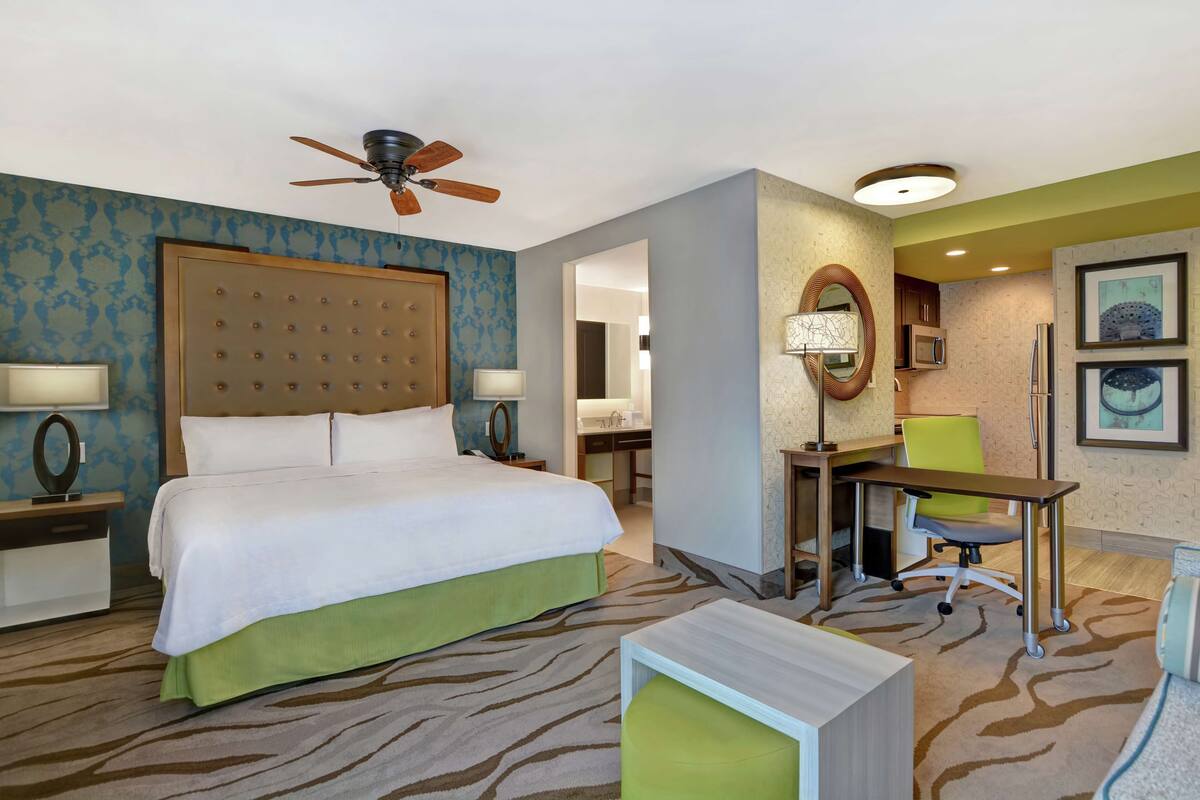 A hotel room with a large bed, a desk, a chair, a ceiling fan, and a bathroom visible through an open door.