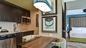 A modern hotel room with a kitchen area, a dining table, striped chairs, a bed with a tufted headboard, and framed artwork on the wall. - Homewood Suites by Hilton Savannah-North/Airport (Savannah)