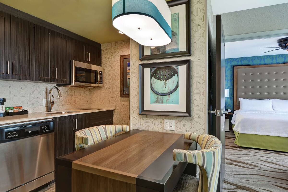 A modern hotel room with a kitchen area, a dining table, striped chairs, a bed with a tufted headboard, and framed artwork on the wall.
