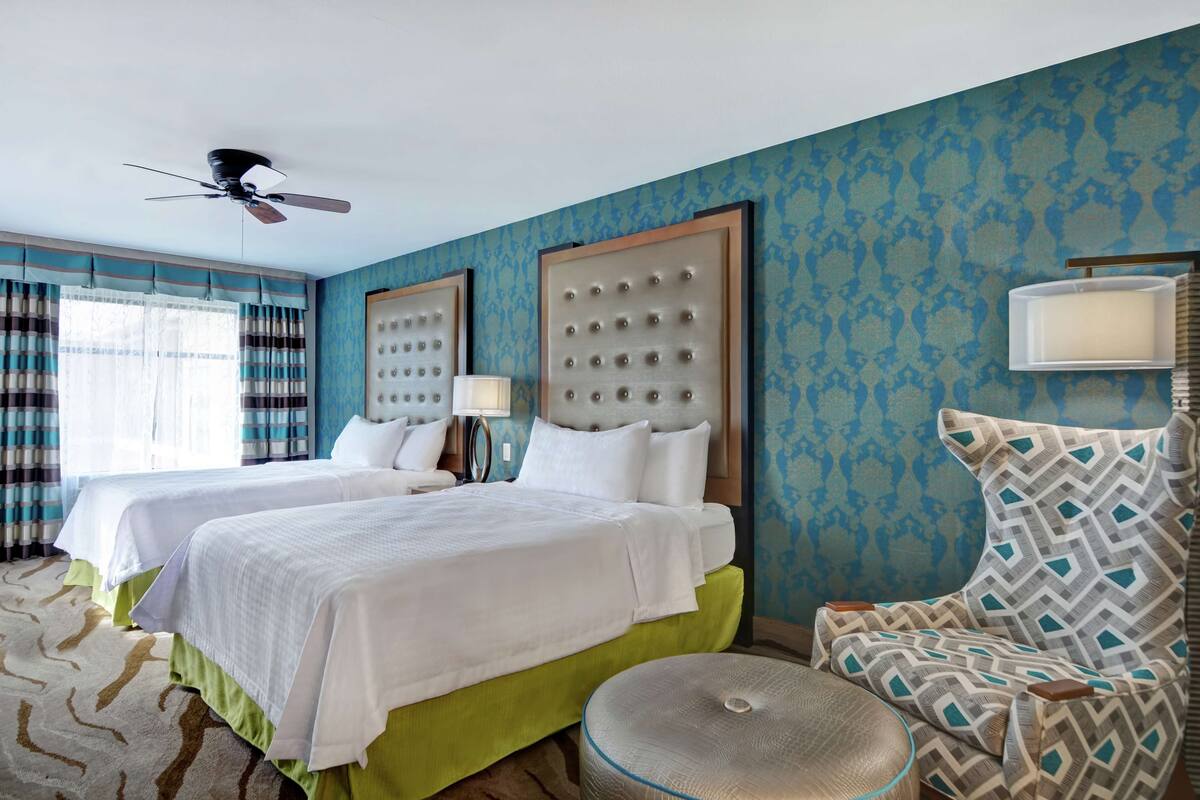 A hotel room with two beds, a ceiling fan, a patterned wall, and a chair with a cushion.