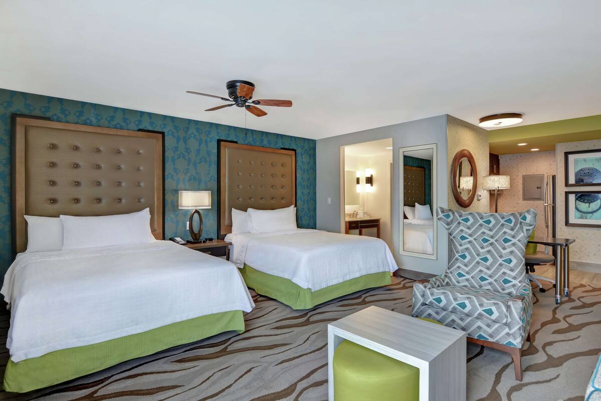 A hotel room with two beds, a ceiling fan, a patterned carpet, a green armchair, and a small table.