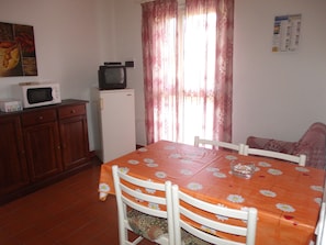 A dining area with a table covered in a floral tablecloth, white chairs, a microwave, a small refrigerator, and a television on top of the fridge. - SVS - Budoni Sa Capannizza (Budoni)